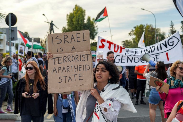 a person holds a sign saying Israel is an apartheid state at a protest with Palestinian flags